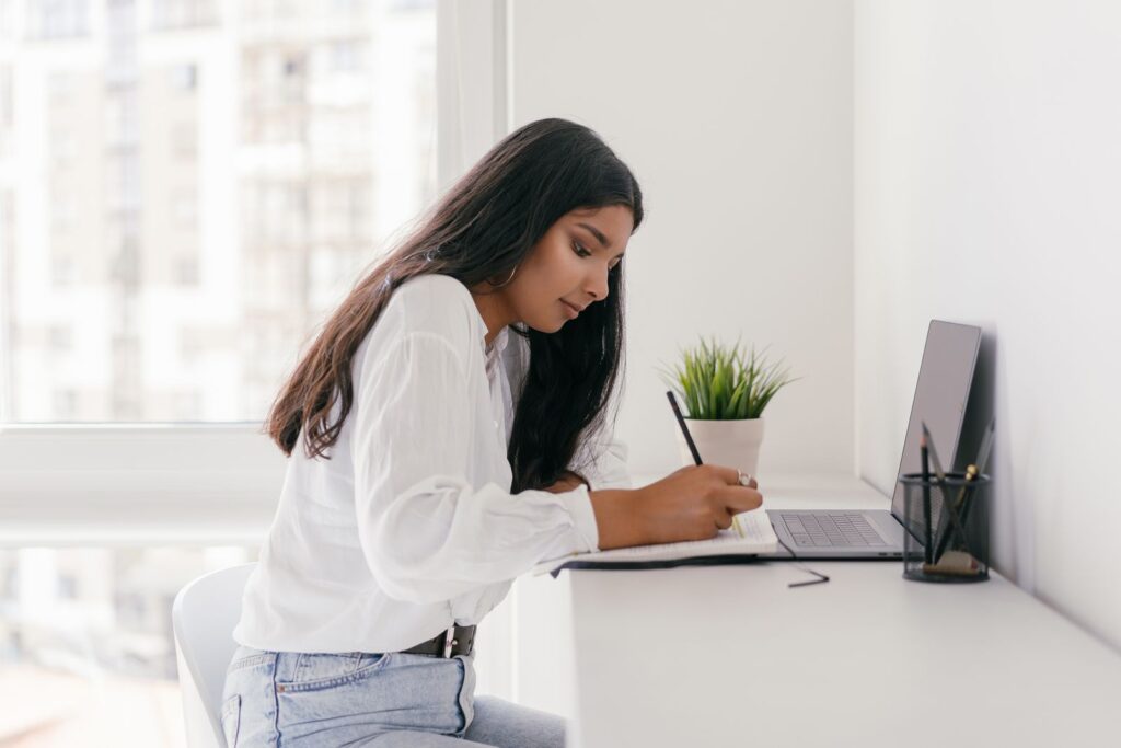 Woman writing in a notebook while using a laptop
